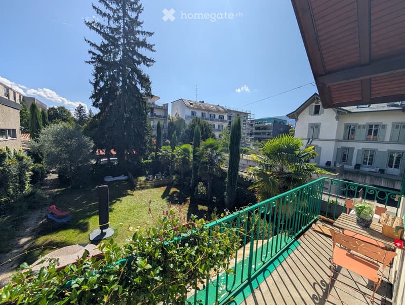 Balcony overlooking a lush garden with tall trees, featuring a green metal railing, a small table, and chairs.