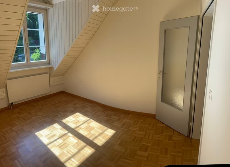 Empty room with wooden flooring, white walls, a window with a view of greenery, and a closed door.