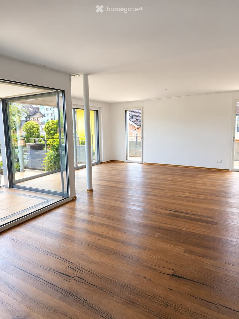 Empty room with wooden flooring, white walls, large glass sliding doors leading to a balcony, and a white column in the center.