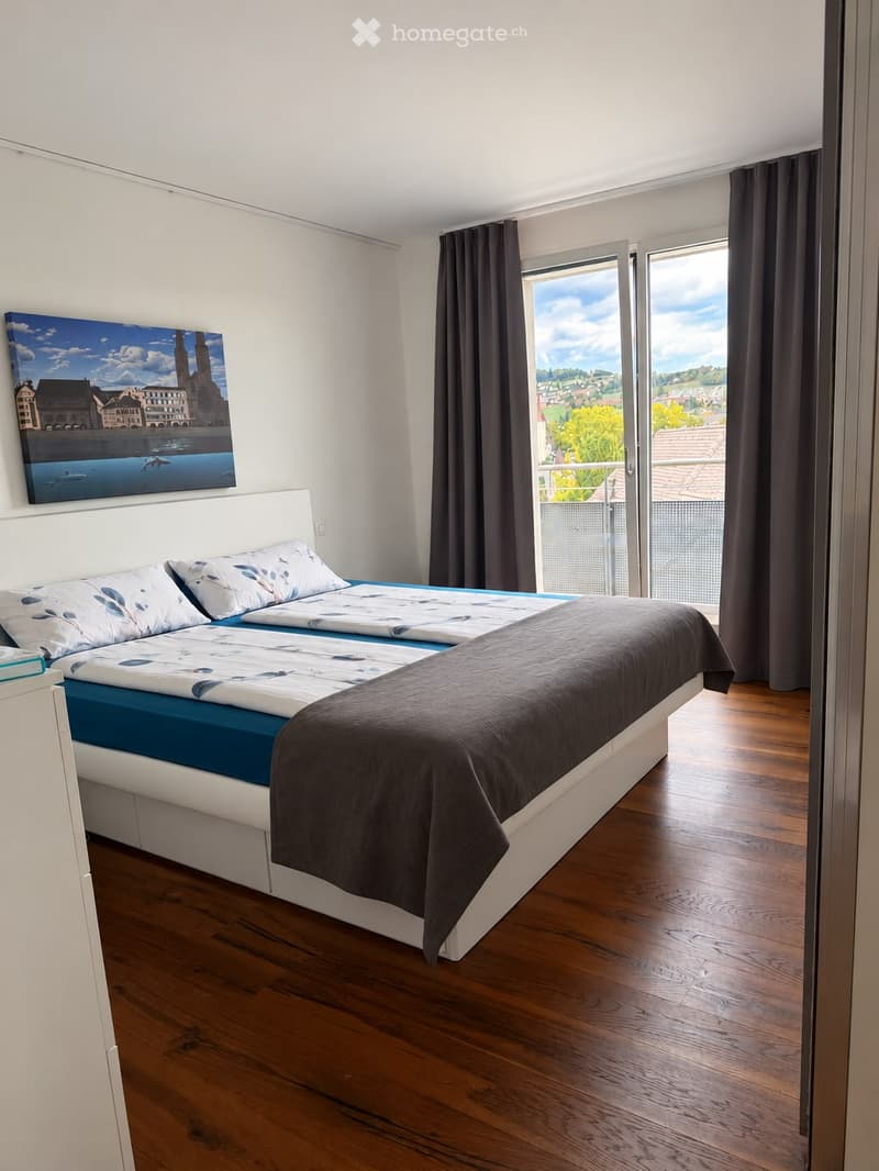 Modern bedroom with a double bed, blue and white bedding, wooden flooring, large window with dark curtains, and a scenic cityscape view.