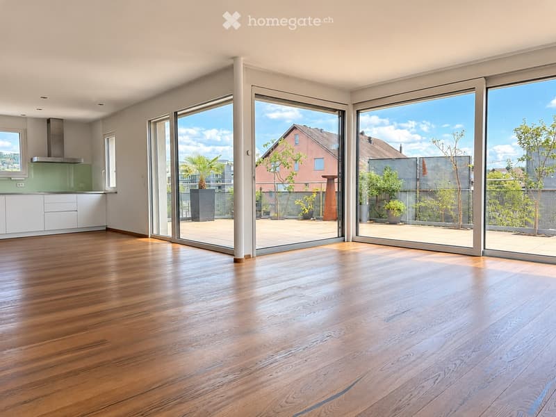 Bright, empty room with wooden flooring, large sliding glass doors leading to a balcony, and a modern kitchen area visible in the background.
