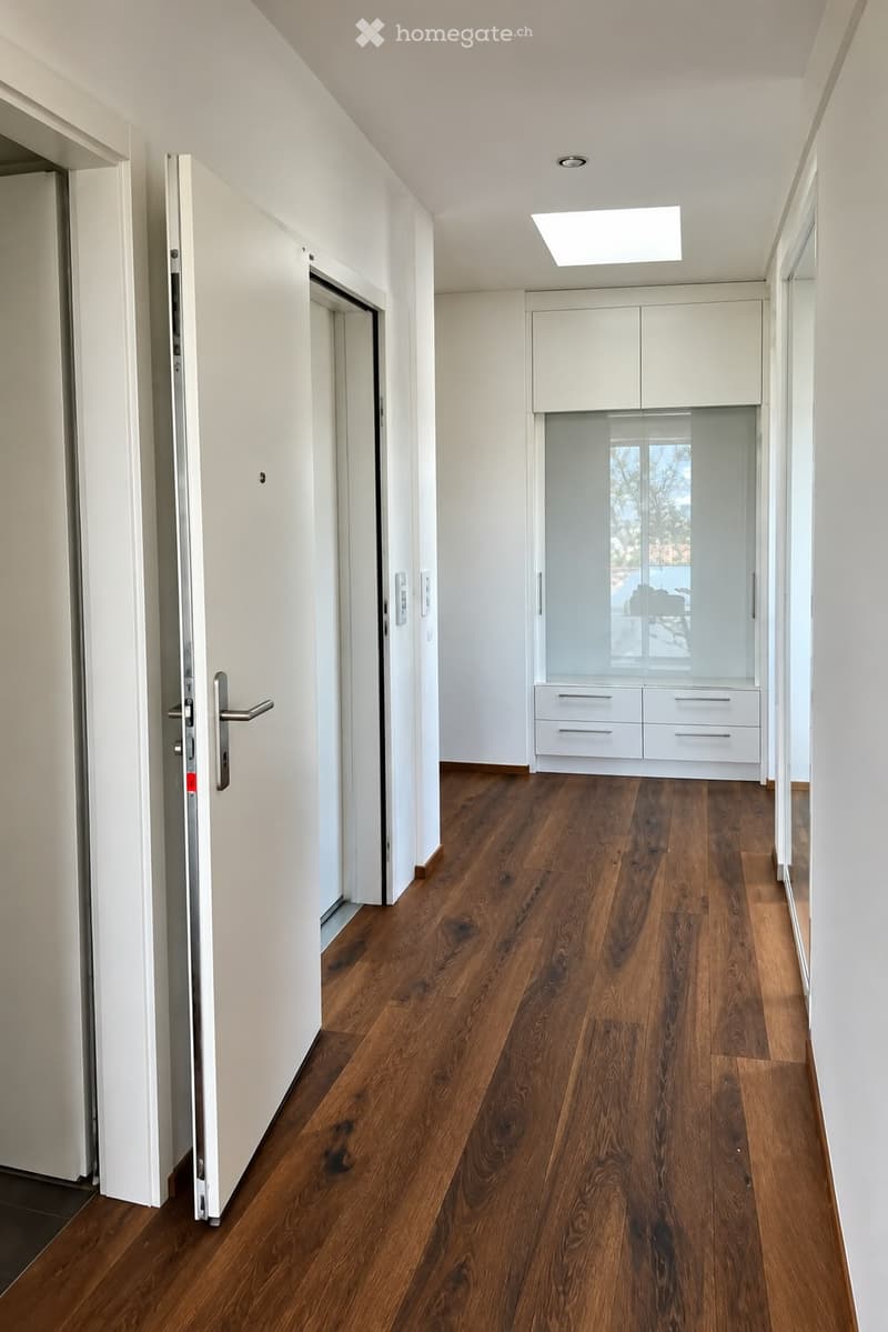 Modern hallway with wooden flooring, white walls, and built-in storage units with glass doors. Features recessed lighting and a partially open door leading to another room.