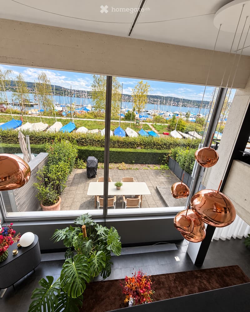 Modern interior with large windows offering a scenic view of a harbor and boats. Features a white dining table with chairs, potted plants, a flower arrangement, and decorative lamps.