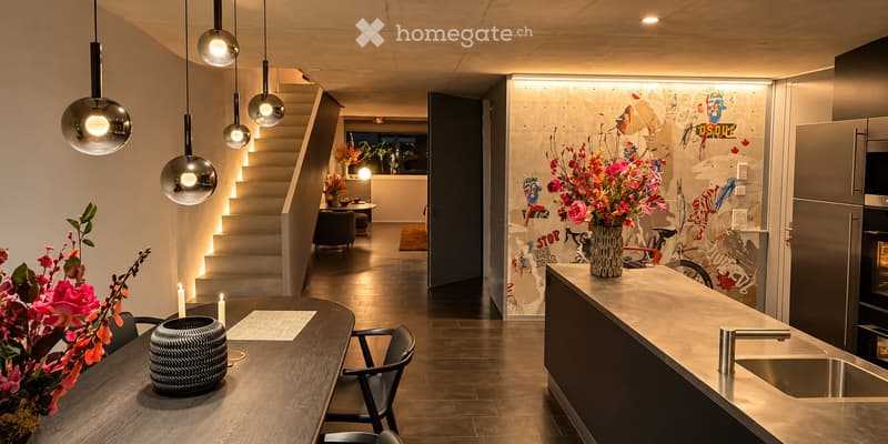 Modern kitchen and dining area with a wooden dining table, chairs, pendant lights, a vase with flowers, and a sink with a faucet.