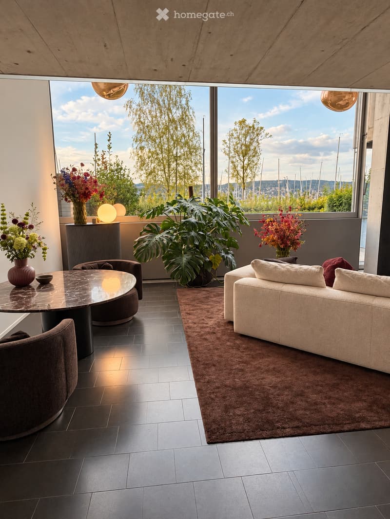 Modern living room with large windows offering a scenic view, featuring a white sofa, coffee table, armchairs, vases with flowers, and a large leafy plant.