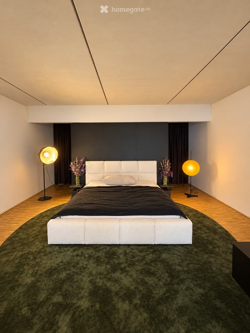 Modern bedroom with a large white upholstered bed, dark green carpet, two nightstands with vases, and floor lamps on either side.