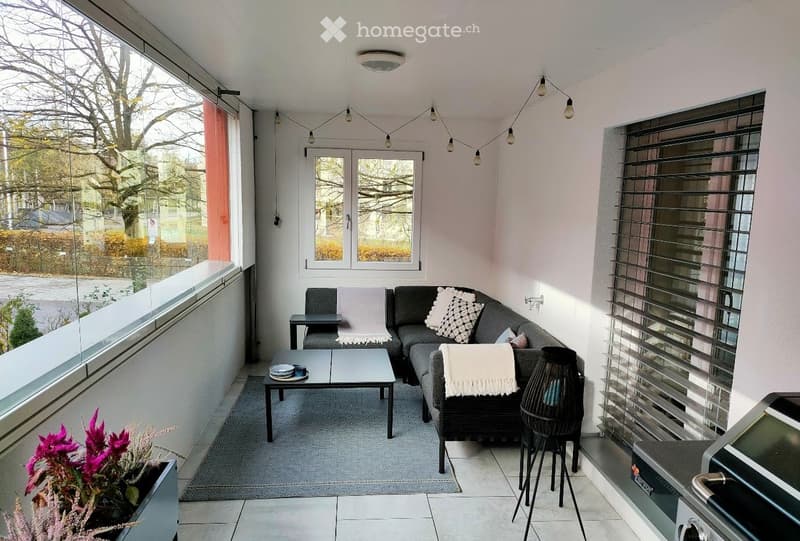 Modern balcony with large windows, grey sofa, coffee table, side table, and decorative pillows.