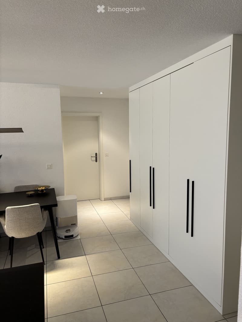 Modern, minimalist room with tiled flooring, white cabinetry, a small table with a chair, and a robotic vacuum cleaner.