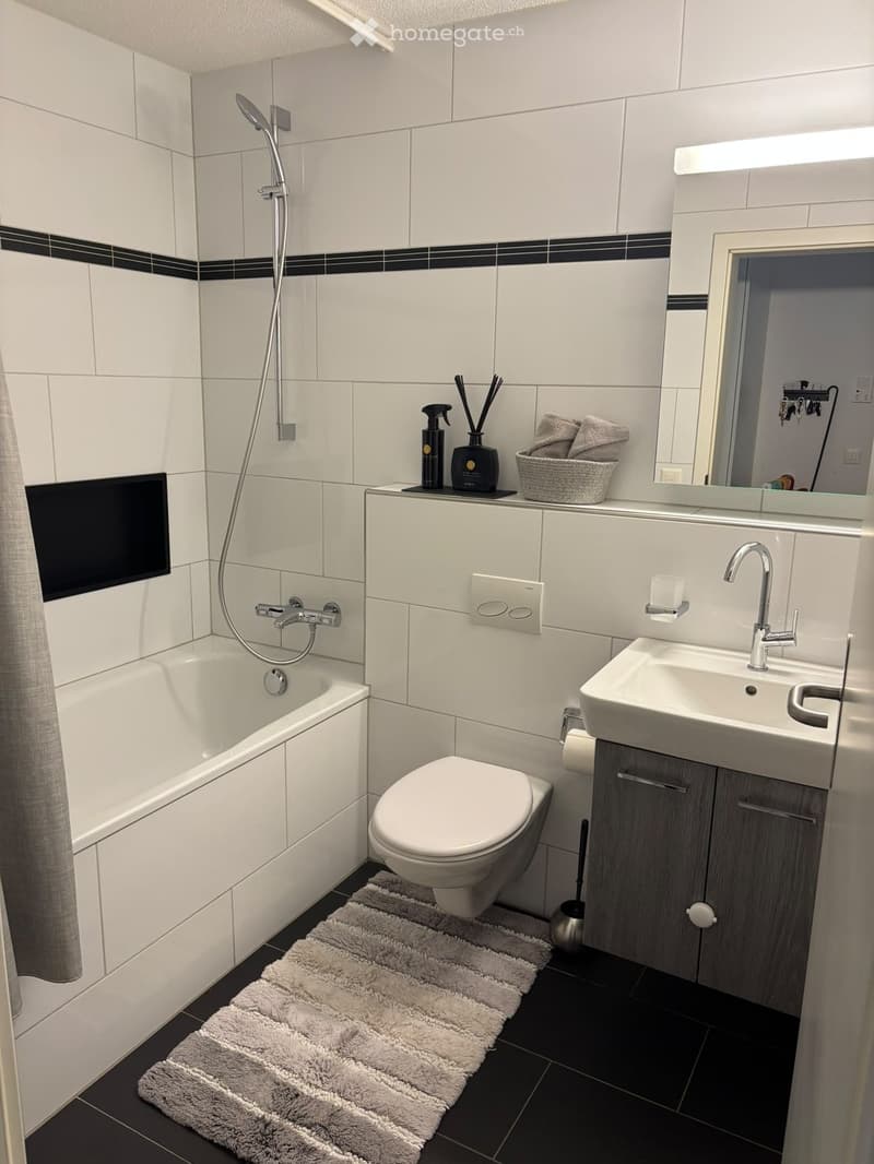 Modern bathroom with white tiles, bathtub, toilet, sink, mirror, and a grey towel on the floor.