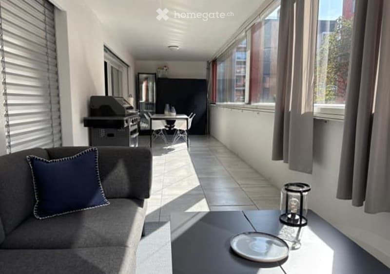 Modern furnished balcony with a grey sofa, coffee table, barbecue, dining table, and chairs. Large windows provide ample natural light.