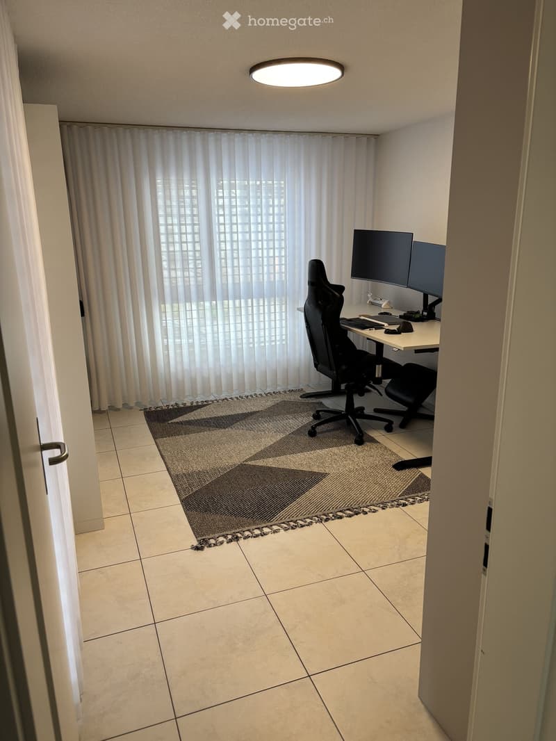 A modern, furnished room with a desk setup, two monitors, a chair, and a large window with curtains.