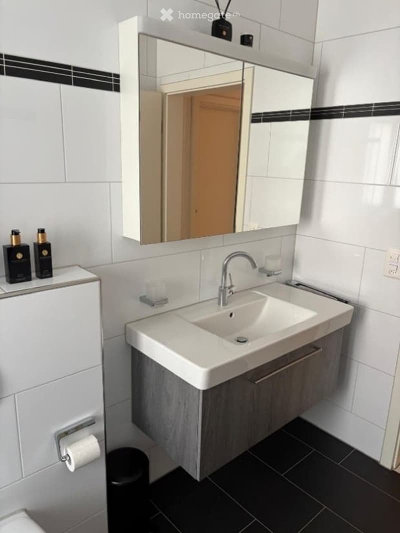 Modern bathroom with white tiles, a large mirror, a sink with a modern faucet, and a small stool. There are decorative candles and a toilet paper holder visible.