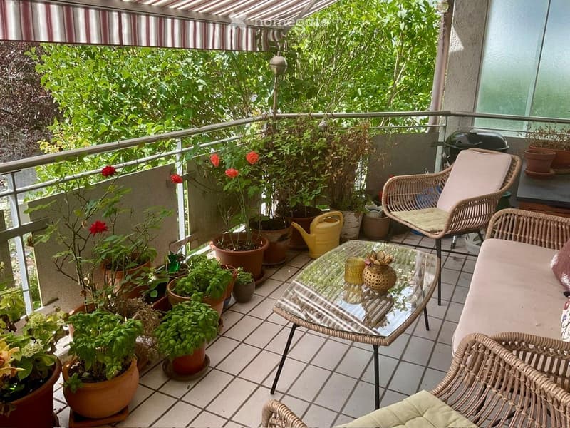 Balcony with tiled floor, metal railing, and striped awning. Features several potted plants, a glass coffee table, wicker chairs, and a small table with decorative items.