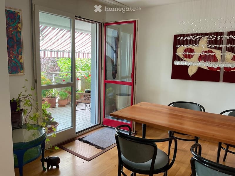 Bright room with wooden floor, large sliding glass doors opening to a balcony, a wooden dining table with chairs, a glass side table, potted plants, and artwork on the wall.