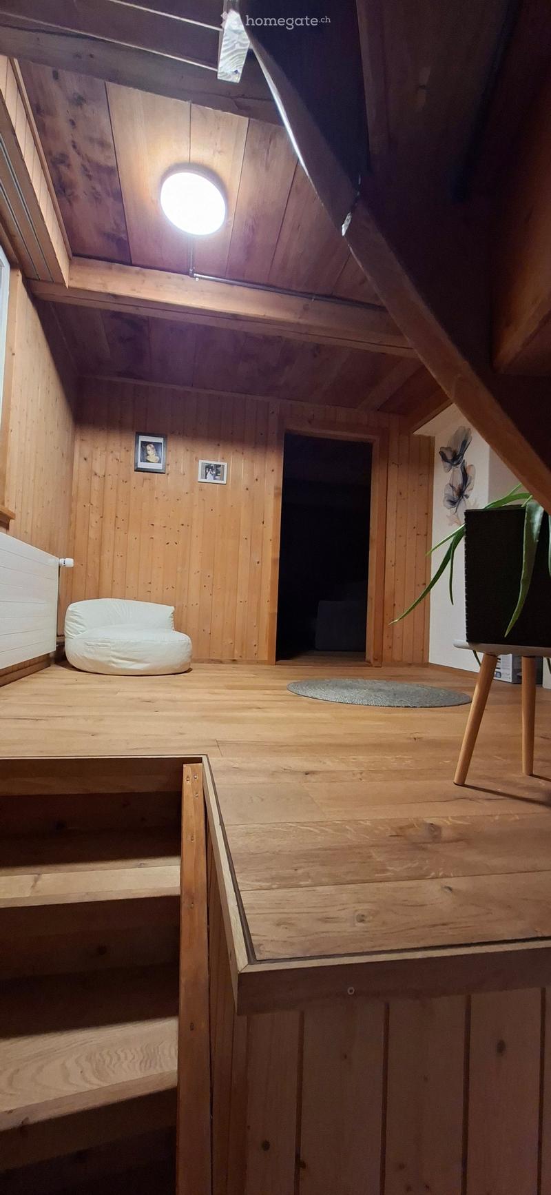 A cozy, wooden-furnished attic room with a small white sofa, a round wooden table with a plant, and a few picture frames on the wall. The room features exposed wooden beams and a skylight.