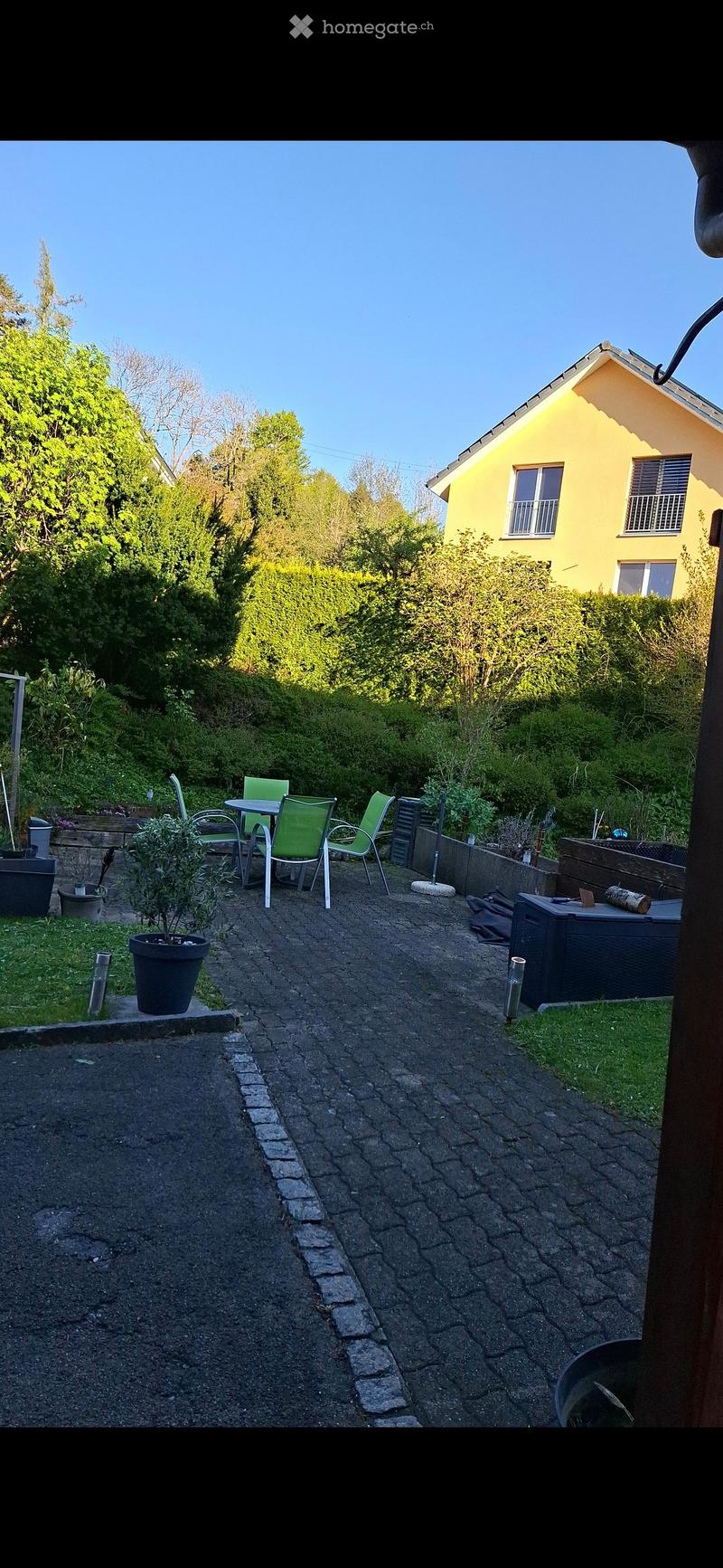 Garden area with a paved pathway, small seating area with a table and chairs, potted plants, and a yellow house in the background.
