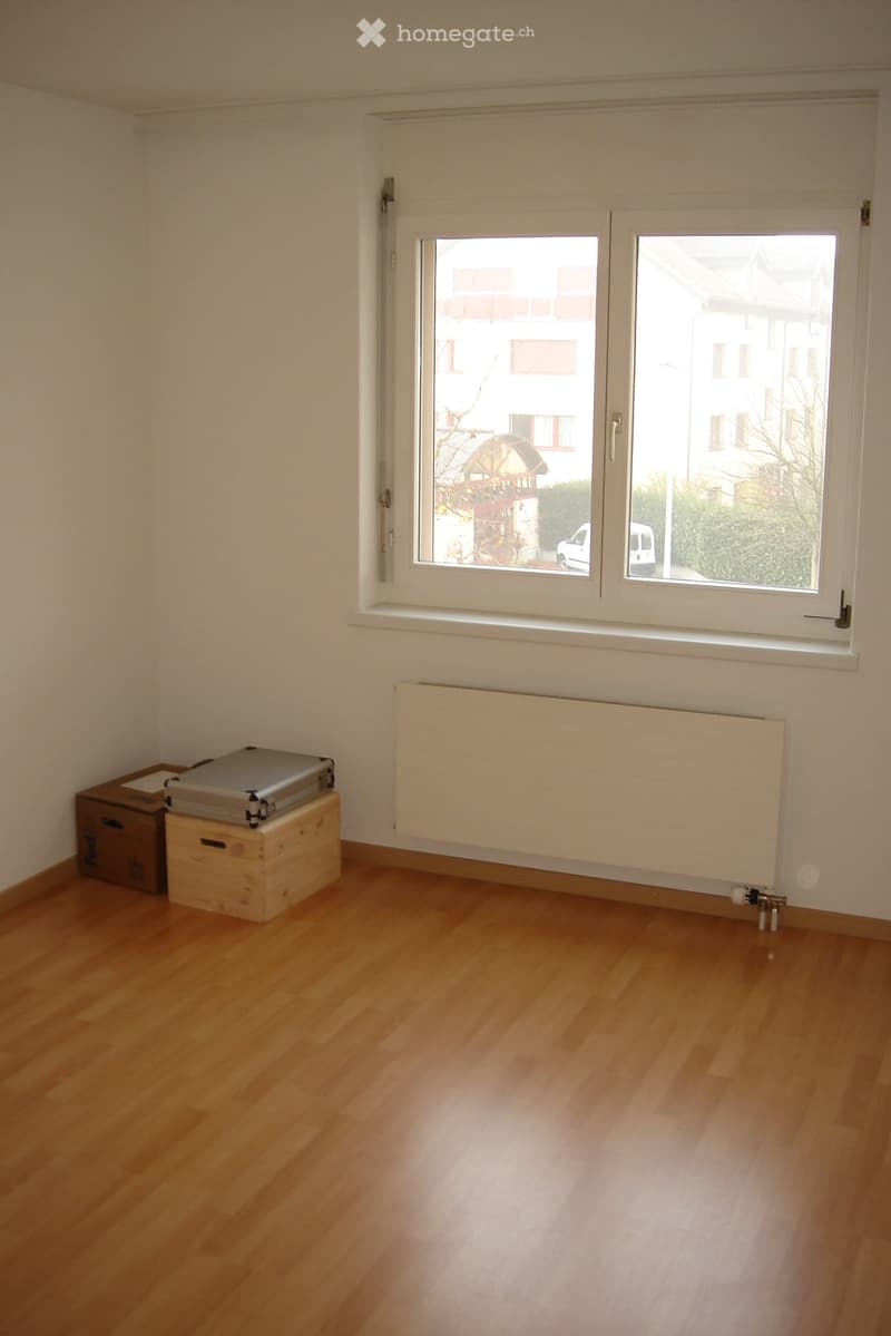 Empty room with wooden flooring, white walls, and a large window with a radiator below it. There are two wooden crates and a cardboard box in the corner.