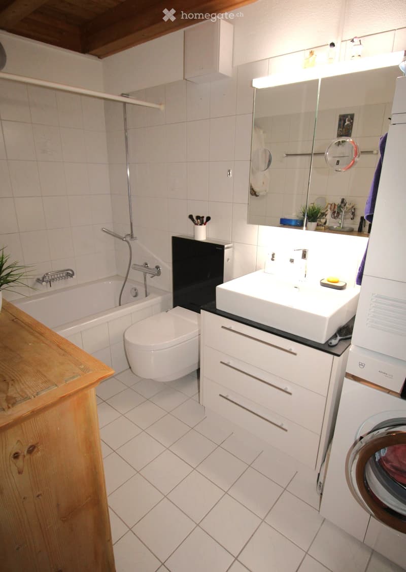 Modern bathroom with white tiles, a bathtub, toilet, sink, mirror, and a washing machine. Features a wooden shelf and a small plant on the counter.