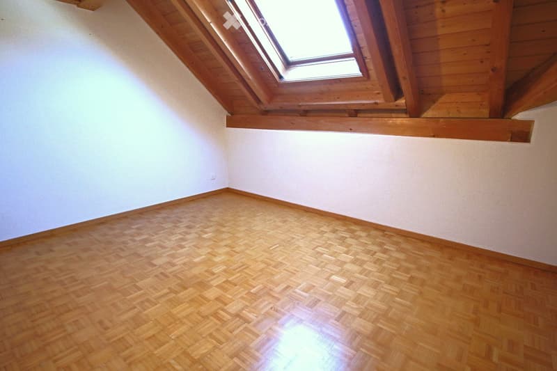 Empty room with wooden herringbone flooring, white walls, and a skylight window in the slanted wooden ceiling.