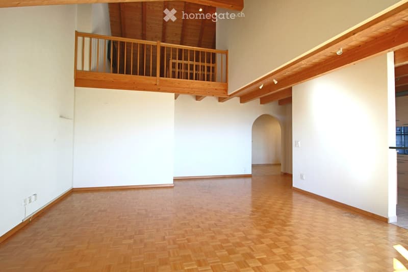 An empty room with wooden flooring and white walls, featuring a wooden balcony level above.