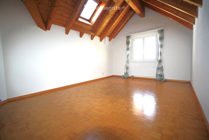 Empty room with wooden flooring, white walls, and exposed wooden beams on the ceiling. Features a window with curtains and a radiator below it.