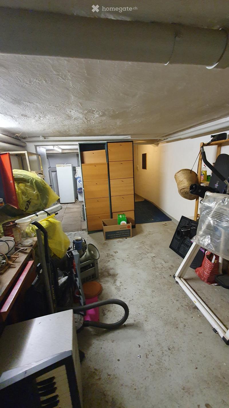 The image shows a cluttered garage or storage area with various items including a refrigerator, cabinets, a vacuum cleaner, a stool, and shelves with miscellaneous objects.