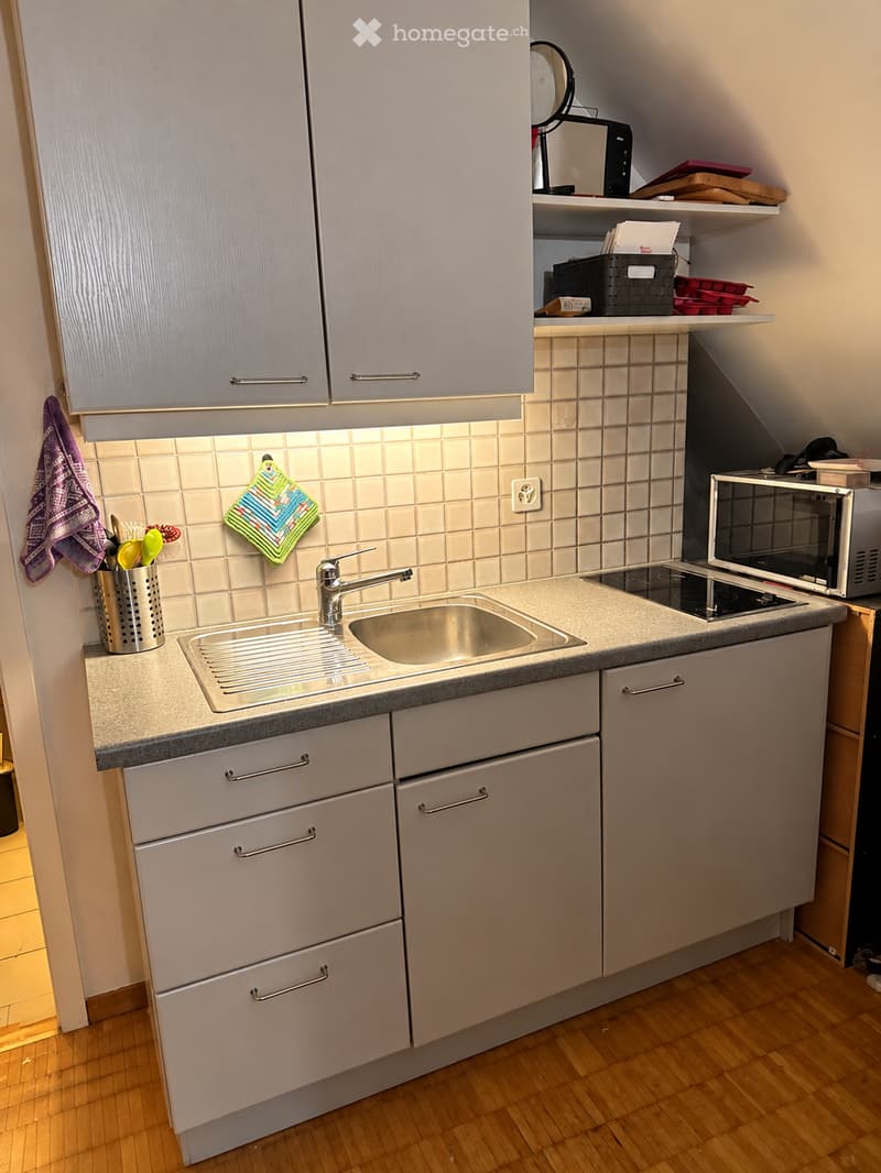 Modern kitchen with light grey cabinets, stainless steel sink, microwave, and wooden flooring.