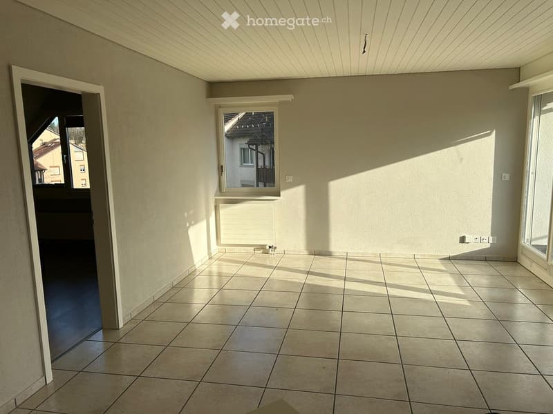 Empty room with tiled floor, white walls, and large windows allowing natural light. There is a door leading to another area and a radiator below one of the windows.