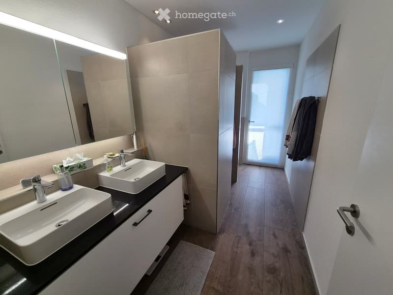 Modern bathroom with two white sinks, sleek faucets, large mirror, and a towel hanging on the wall. The space features light-colored tiles and a wooden floor.