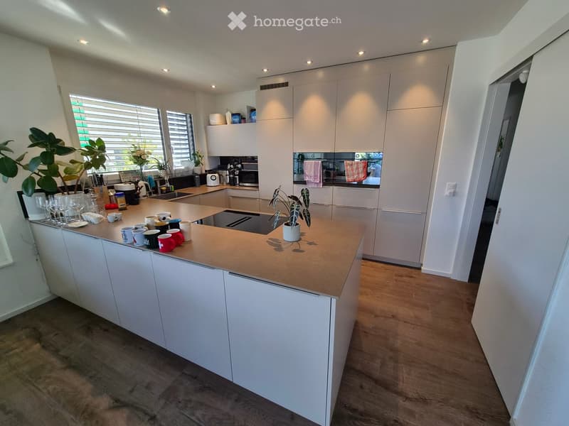 Modern kitchen with white cabinets, a central island with a wooden countertop, and various appliances including a sink, oven, microwave, toaster, and coffee maker. The island is topped with cups, bowls, wine glasses, and a small plant. The kitchen feature