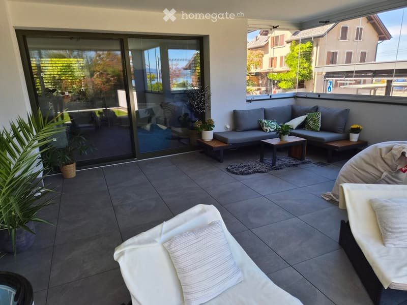 Modern balcony with grey sofa, small wooden table, white lounge chairs, and potted plants, offering a scenic view of the surroundings.