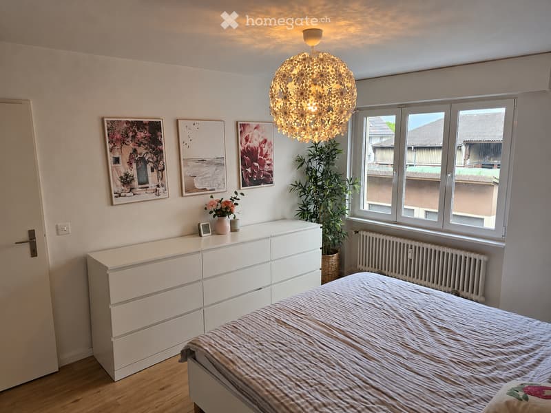 Bright bedroom with a bed, white dresser, decorative picture frames, a potted plant, and a modern light fixture.