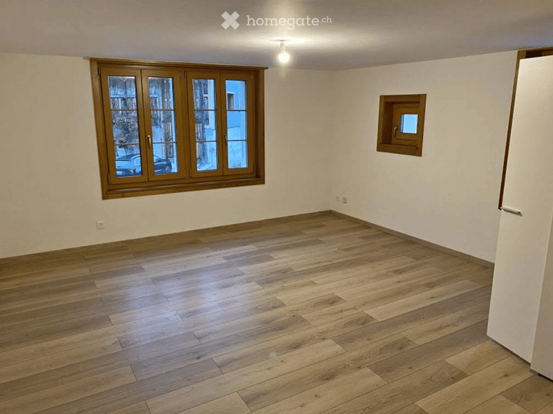 Empty room with wooden flooring, large window with wooden frame, small window, and white walls.