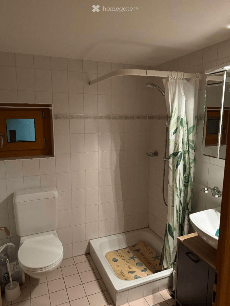 Bathroom with tiled walls and floor, featuring a toilet, bathtub with a decorative bath mat, a sink with a wooden cabinet, and a shower area with a curtain.