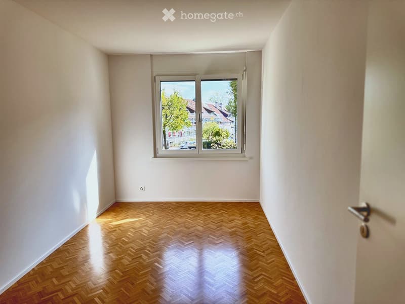 Empty room with wooden herringbone flooring, white walls, and a large window with a view of the street and trees.