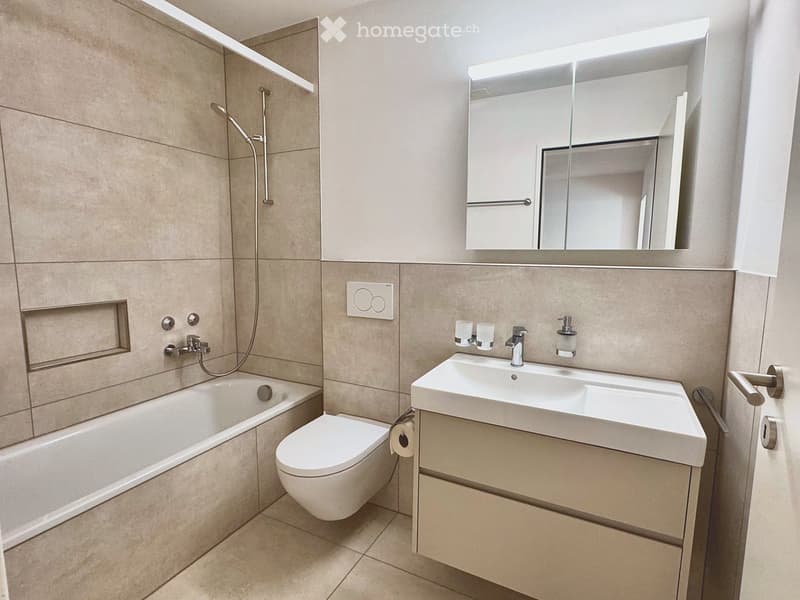 Modern bathroom with beige tiles, a bathtub, toilet, sink, mirror, and showerhead.