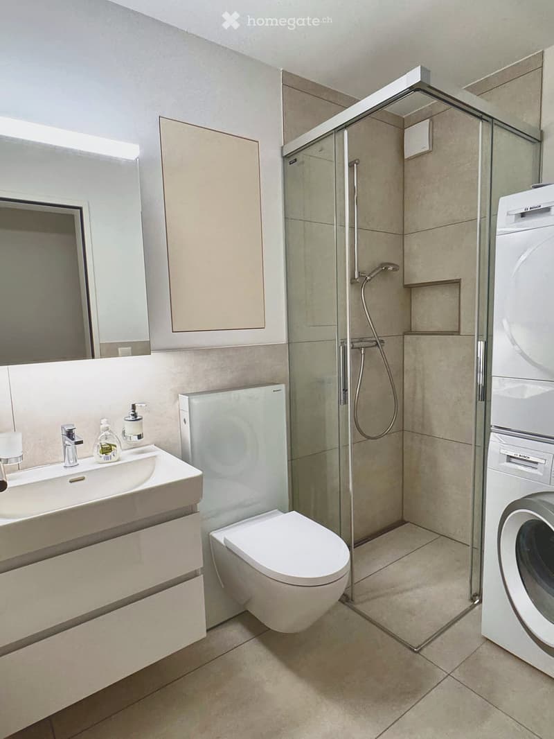 Modern bathroom with a white sink, toilet, glass-enclosed shower, and a Bosch washing machine. Features a large mirror and a sleek design with beige tiles.