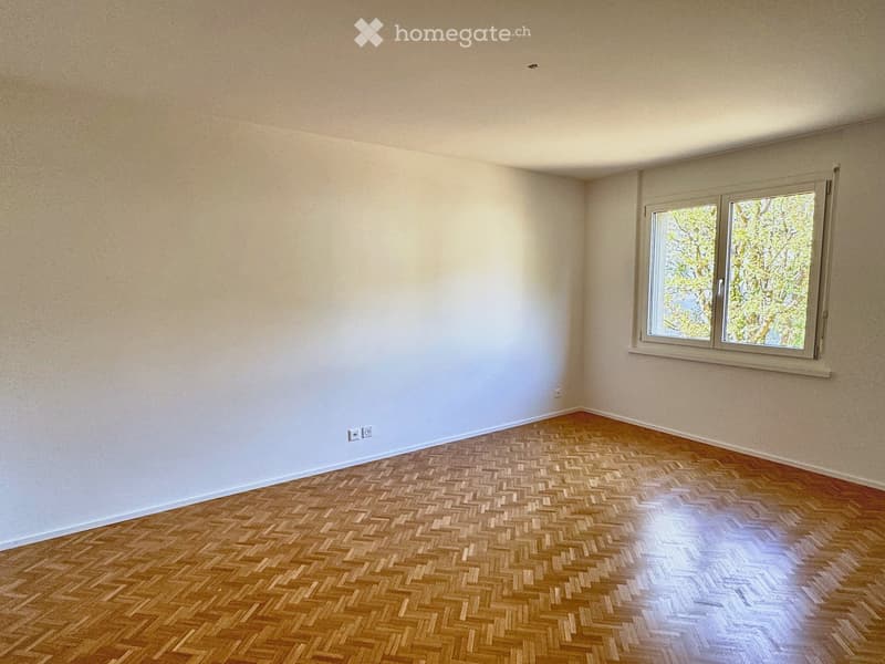 Empty room with wooden herringbone flooring and white walls, featuring a large window with a view of greenery.