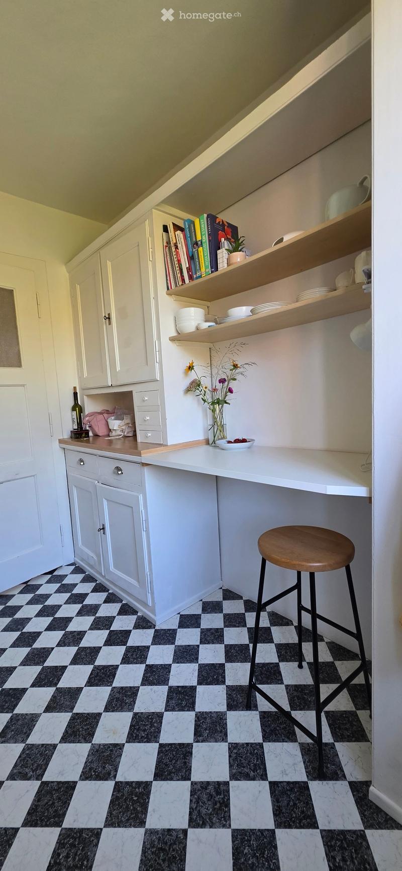 A compact kitchen area with white cabinets, wooden shelves, a white countertop, and a black and white checkered floor. A wooden stool is placed near the counter, and various items such as a bottle, vase, bowl, and books are visible on the shelves and coun