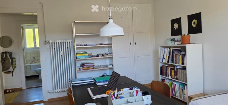 A well-organized home office with a wooden desk, laptop, chair, bookshelf filled with books, radiator, mirror, potted plant, framed picture, calendar, and various office supplies.