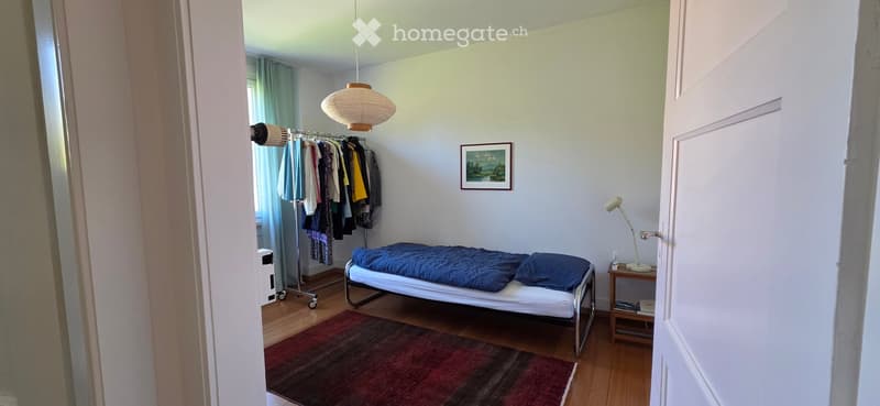 A small bedroom with a single bed, nightstand, lamp, picture frame, clothing rack, and fan. The room has wooden flooring and a colorful rug.