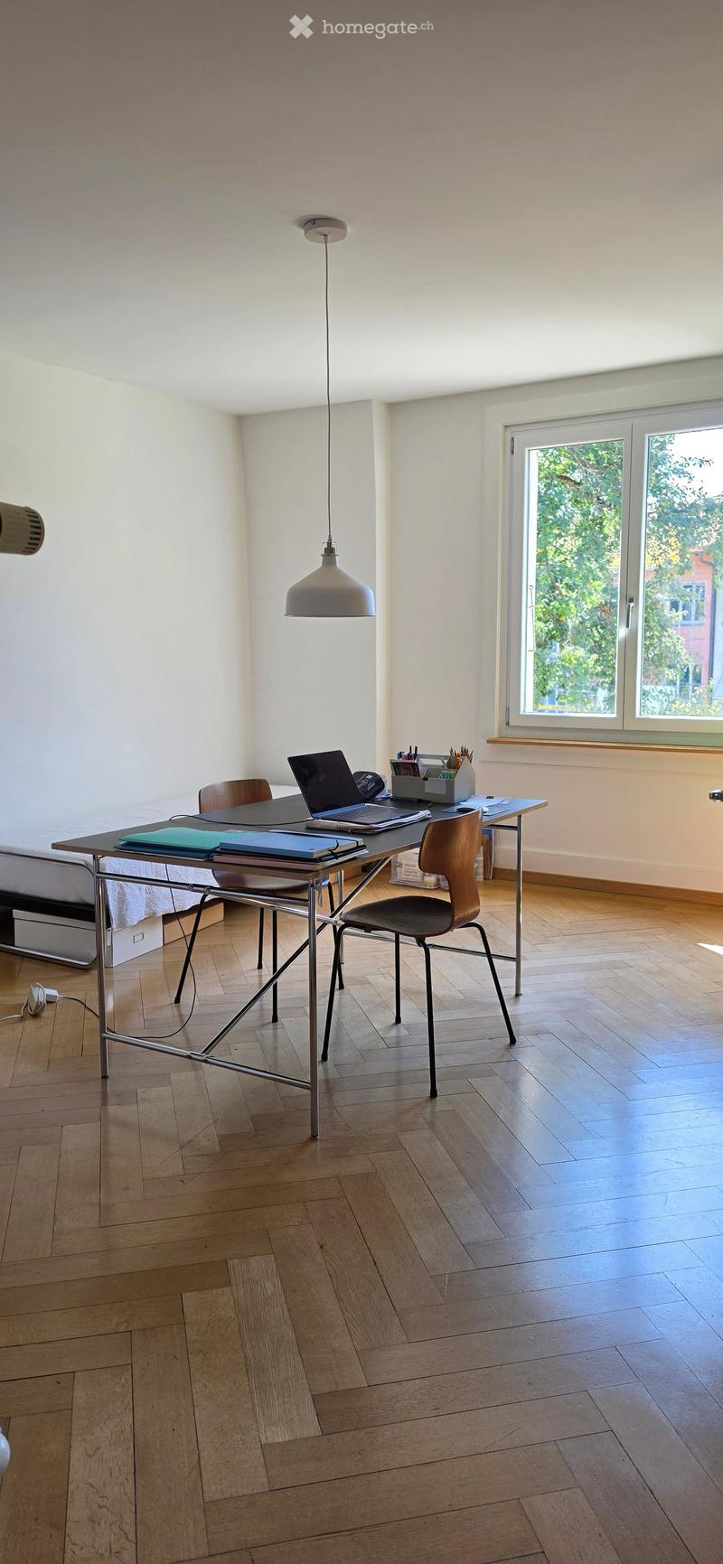 A well-lit room with wooden flooring, a modern desk setup including a laptop, books, and a pen holder, accompanied by a hanging lamp and a comfortable chair.