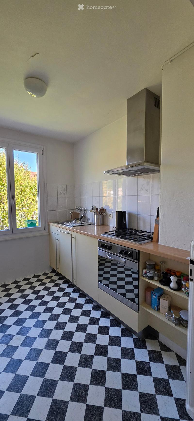 The kitchen features a checkered black and white tile floor, white cabinets, a wooden countertop, and stainless steel appliances including a stove, oven, and sink. A window with a view of greenery is visible on the left side.