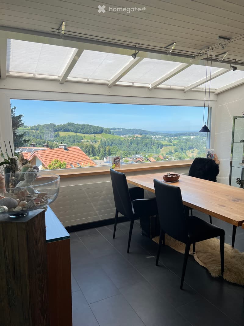 Bright dining area with large windows offering scenic views, featuring a wooden table, black chairs, a decorative bowl, and a glass vase with seashells.