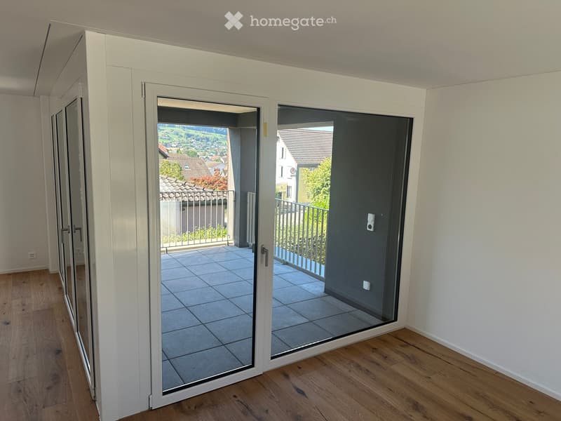 View of a modern interior with large glass sliding doors leading to a tiled balcony area, overlooking a residential neighborhood with visible houses and greenery.