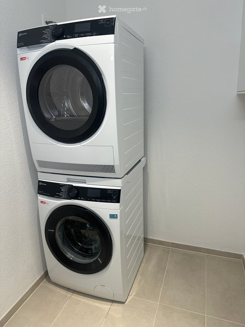 A modern, white washing machine and dryer set is positioned against a light grey wall with a beige tiled floor.