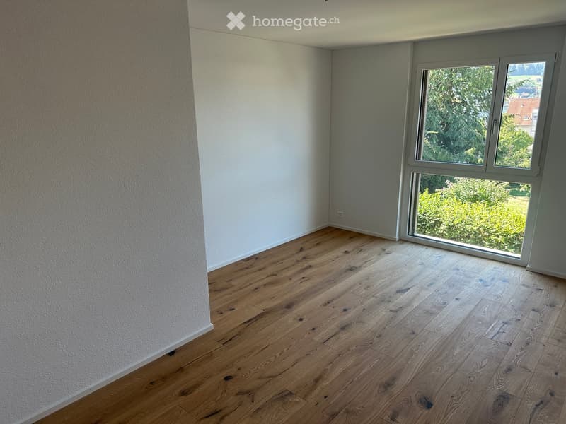 Empty room with light wooden flooring and white walls, featuring a large window with a view of greenery outside.