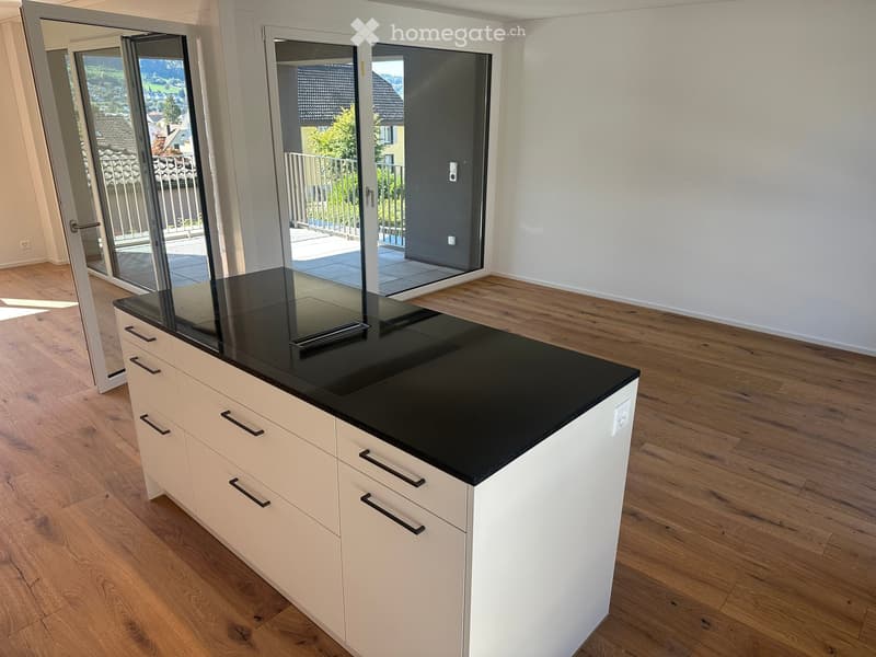 Modern kitchen with a white cabinet island featuring a black countertop, wooden flooring, and large glass doors leading to a balcony.