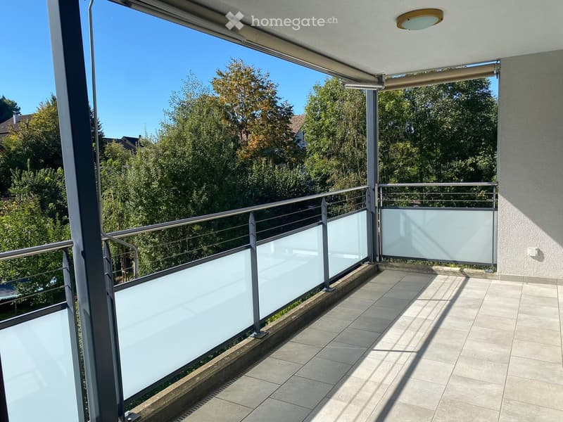 Balcony with modern glass balustrade, tiled floor, and retractable awning, overlooking a green area with trees.