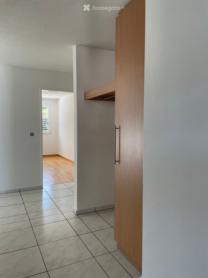 Empty hallway with tiled floor, wooden closet door, and adjacent room with wooden flooring and window.